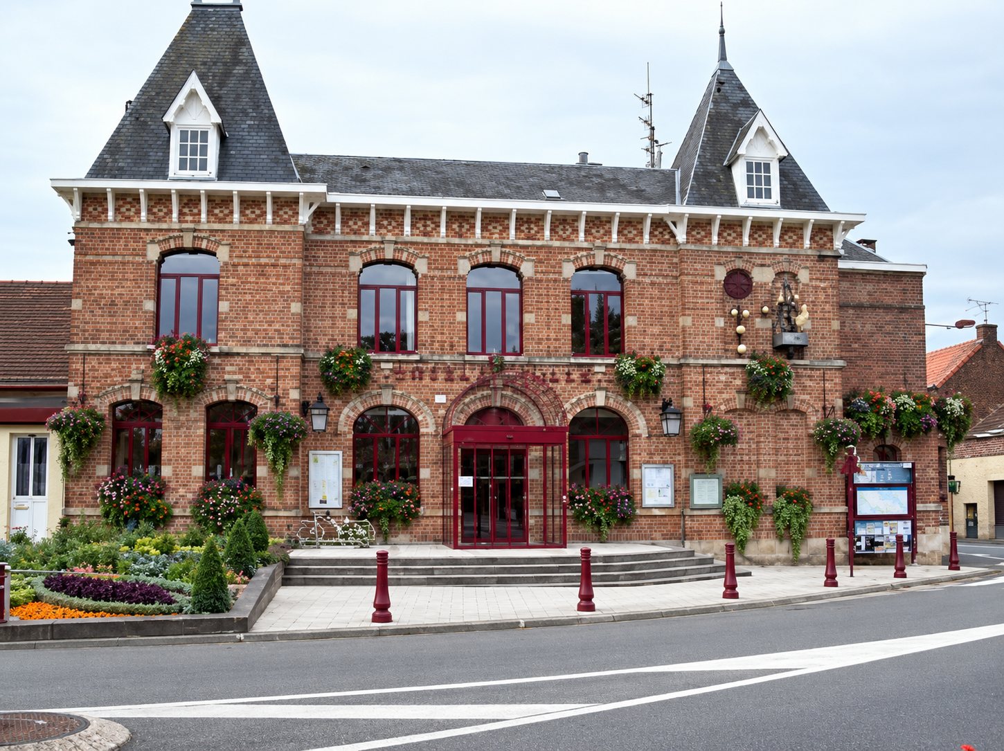 Façade d’un bel édifice en briques rouges à toit d’ardoise, orné de jardinières fleuries, avec un escalier d’entrée et des bornes rouges devant.