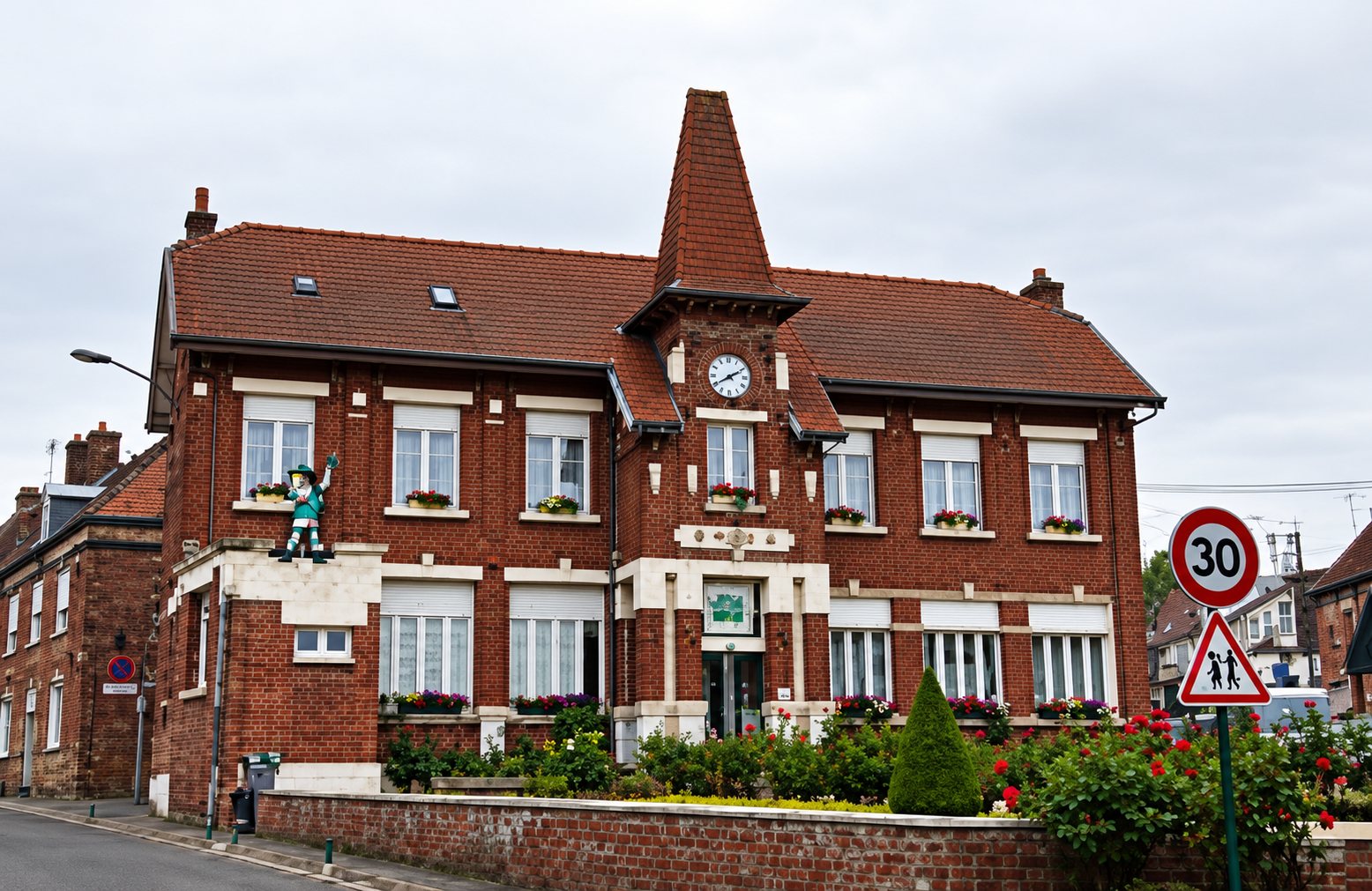 Un grand bâtiment en briques rouges à toit pointu avec une horloge, des fenêtres fleuries et un panneau de limitation de vitesse à 30 au premier plan.